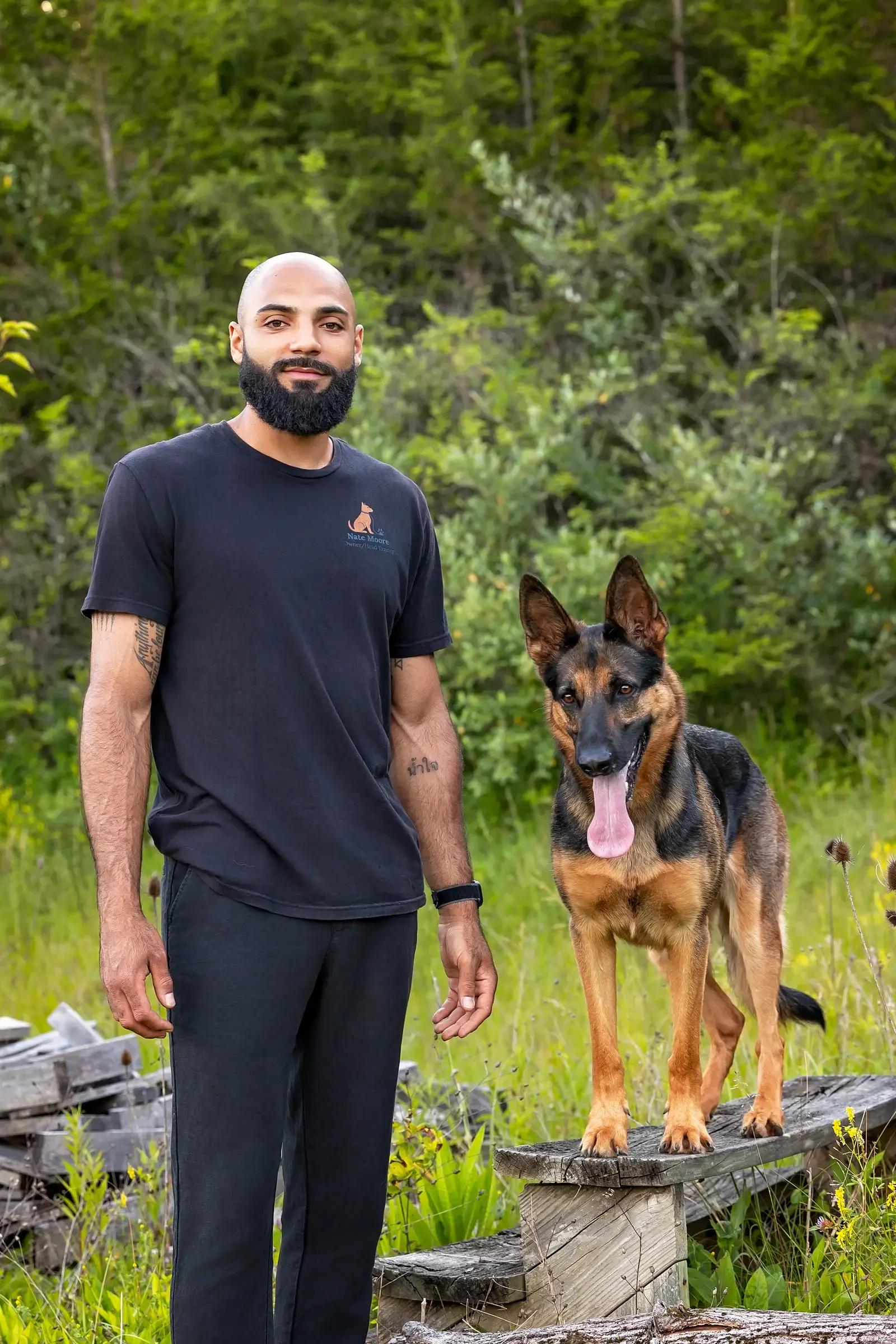 Nate Moore standing in a field beside a German Shepherd, wearing a Moore Canine Co. trainer tee